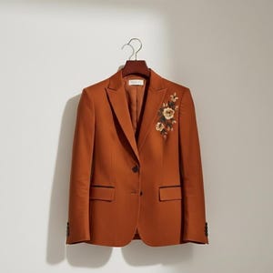 May include: A burnt orange blazer with floral embroidery on the upper left. It features a notched lapel, two front pockets, and a single-button closure. The blazer is suspended on a wooden hanger.