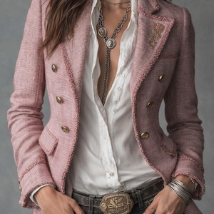 May include: A pink tweed blazer with gold buttons and a white collared shirt. The blazer has a decorative floral detail on the lapel. The outfit includes a silver necklace, a belt with a decorative buckle, and gray jeans.