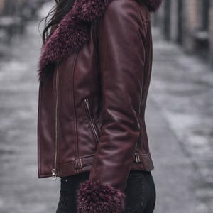 May include: Burgundy leather jacket with a faux fur collar and cuffs. The jacket features a zipper closure and a tailored fit. The model is wearing black jeans. The jacket is a stylish outerwear option.