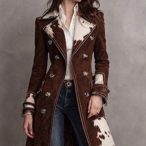 May include: A brown suede coat with white cowhide accents on the collar, cuffs, and lower front. The coat features silver buttons, a matching belt, and white stitching. The model is wearing blue jeans and a white shirt.