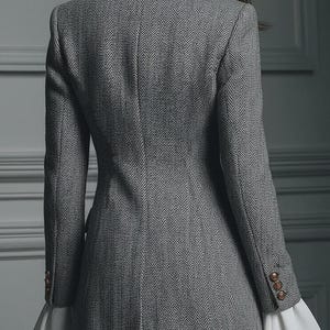 Puede incluir: Un blazer gris con dise&ntilde;o de espiga y botones dorados en las mangas. El blazer tiene un corte entallado y pu&ntilde;os blancos con volantes. La vista trasera muestra el dise&ntilde;o del blazer, adecuado para vestimenta formal o de negocios.