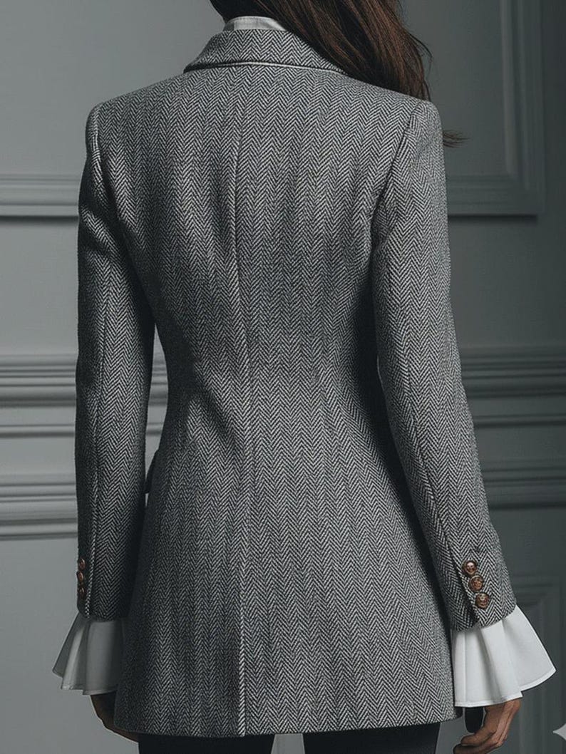 Puede incluir: Un blazer gris con dise&ntilde;o de espiga y botones dorados en las mangas. El blazer tiene un corte entallado y pu&ntilde;os blancos con volantes. La vista trasera muestra el dise&ntilde;o del blazer, adecuado para vestimenta formal o de negocios.