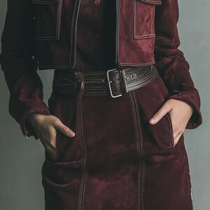 May include: A burgundy suede jacket with two front pockets and a matching skirt. The outfit includes a brown leather belt with a silver buckle. White stitching details the seams of the jacket and skirt. The model has their hands in the skirt pockets.
