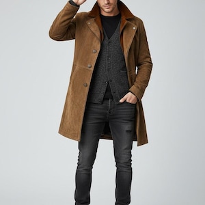 Op de afbeelding: Een bruine suède jas met een contrasterende kraag, gedragen over een donkergrijs vest en een zwart shirt met lange mouwen. De outfit wordt gecompleteerd met een zwarte jeans met distress, een bruine fedora en bruine laarzen. De algehele stijl is klassiek en stijlvol.