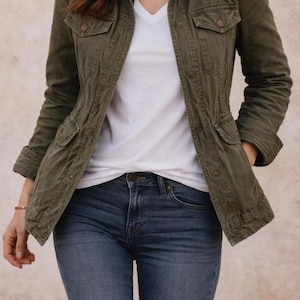 May include: An olive green utility jacket with multiple pockets and button fastenings, worn with a white V-neck top and dark wash blue jeans. The jacket has a classic, casual style.
