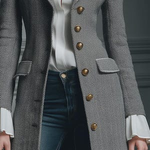Puede incluir: Blazer gris con estampado de espiga, botones dorados y ribete blanco. El blazer entallado tiene dos bolsillos delanteros y se combina con una blusa blanca y vaqueros azules. La blusa blanca tiene mangas con volantes. La chaqueta mide unos 76 cm de largo.