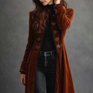 May include: A long, rust-coloured velvet coat with black trim and ornate buttons. The coat is buttoned up and worn over a black turtleneck and dark wash jeans. The coat has a classic, tailored design.