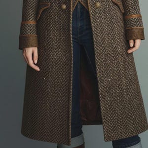 May include: A brown herringbone-patterned coat with gold buttons and brown trim. The coat has a tailored fit and a long length, reaching below the knees. The sleeves have decorative cuffs. The coat is paired with blue jeans and brown boots.