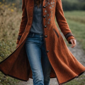 Può includere: Un cappotto lungo color ruggine con un motivo floreale ricamato sul davanti. Il cappotto ha un bordo e bottoni marrone scuro. La persona indossa jeans blu. Il cappotto è un capo di abbigliamento esterno elegante.