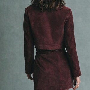 May include: A burgundy suede jacket and matching mini skirt. The jacket is cropped, with long sleeves and a structured fit. The skirt has a wrap design. The outfit is a deep, rich color, and the model is viewed from the back.