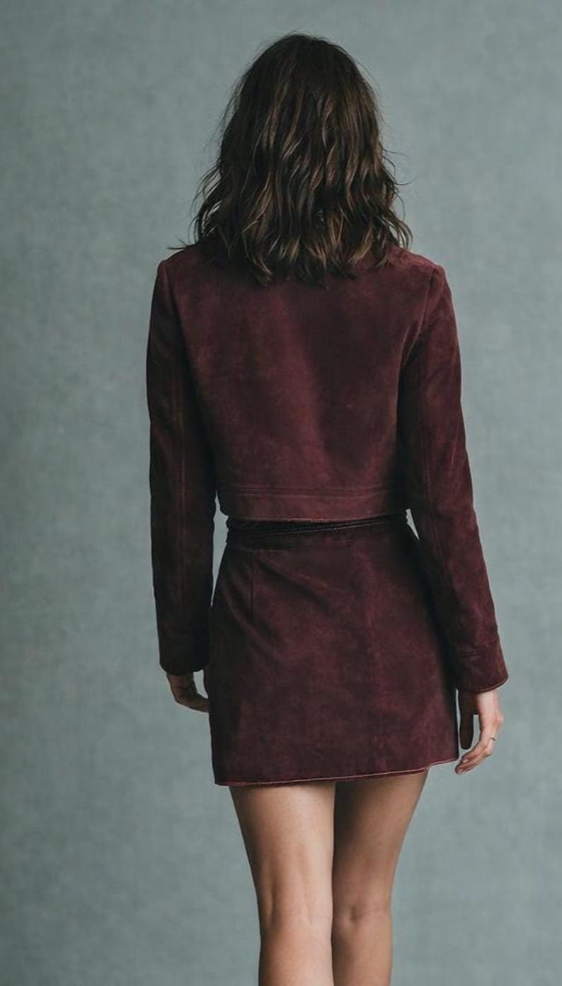 May include: A burgundy suede jacket and matching mini skirt. The jacket is cropped, with long sleeves and a structured fit. The skirt has a wrap design. The outfit is a deep, rich color, and the model is viewed from the back.