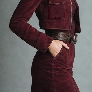 May include: A burgundy suede jacket with two front pockets and a matching mini skirt. The jacket has a zipper closure and the outfit is accessorized with a brown leather belt. The outfit is a stylish ensemble.