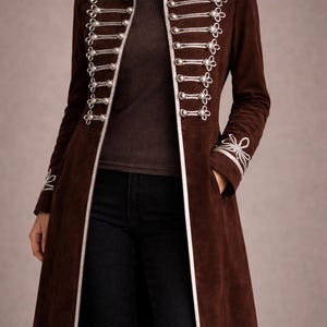 May include: A long, brown suede coat with silver trim and decorative buttons. The coat features a high collar and detailed embellishments on the sleeves and shoulders. The coat is worn over a brown top and black trousers, creating a stylish, layered look.