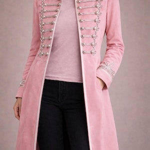 Può includere: Un cappotto lungo rosa con abbellimenti e bottoni color argento. Il cappotto presenta un colletto alto e finiture decorative sulle spalle e sui polsini. Indossato sopra un top rosa chiaro e pantaloni neri.