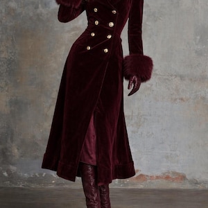May include: A long, burgundy velvet coat with a double-breasted design and gold buttons. The coat features a faux fur collar and cuffs, and is paired with matching gloves and boots. The coat is floor-length and has a high slit.