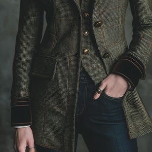 Pu&ograve; includere: Un blazer in tweed verde e marrone con gilet abbinato e bottoni dorati. Il blazer ha una vestibilit&agrave; sartoriale, con polsini in velluto marrone scuro e una tasca. La persona indossa jeans blu scuro e anelli.