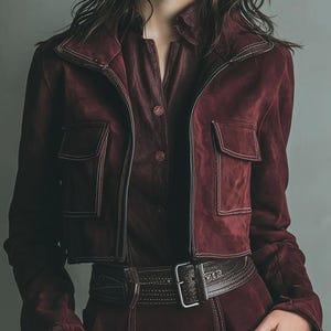 May include: A burgundy suede jacket with two front pockets and a matching burgundy shirt. The jacket has a zipper closure and a collar. A brown belt with a silver buckle cinches the waist. The outfit is a monochromatic ensemble.