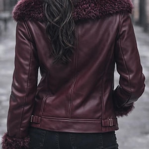 May include: Burgundy leather jacket with a faux fur collar and cuffs. The jacket features a classic design with a tailored fit, visible stitching, and buckle details at the waist. The model is wearing black jeans.