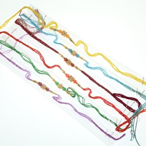 May include: A set of five decorative bracelets featuring multi-coloured threads in yellow, blue, red, green, and purple. Each bracelet includes a central gold-toned metal charm with embedded clear stones and small coloured beads on either side.