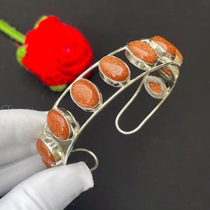 May include: A silver-toned cuff bracelet featuring a series of oval-shaped, shimmering copper-colored goldstone cabochons set in metal bezels. The bracelet has an open-ended design, showcasing a polished metallic frame that holds the stones.
