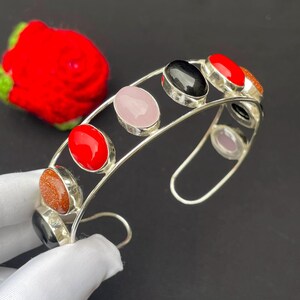 May include: A silver cuff bracelet featuring a double-wire band set with multiple oval-shaped cabochon stones. The stones include polished black, bright red, pale pink, and shimmering gold-flecked brown gems arranged along the open metal frame.