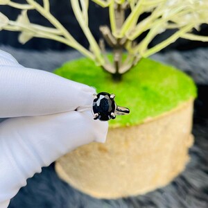 May include: A silver-toned ring featuring a single oval-cut black gemstone held in a four-prong setting. The band is smooth and metallic, showcasing a polished finish that reflects light. The dark stone is centered, creating a contrast with the metal.