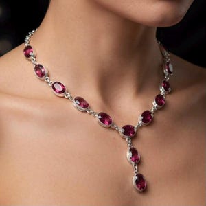 Op de afbeelding: Een zilveren ketting met een reeks ovaalvormige, diep magentakleurige edelstenen in metalen zettingen, met een centraal hangend gedeelte bestaande uit twee extra ovale edelstenen die verticaal aan de ketting hangen.