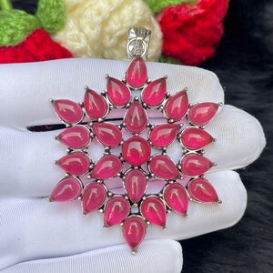 Può includere: Un ciondolo in argento con una pietra preziosa rosa circolare centrale circondata da due anelli concentrici di pietre rosa a forma di goccia disposte a stella, con un gancio in argento sulla parte superiore per una catenina.