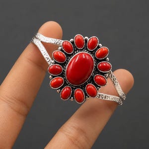 Puede incluir: Una pulsera de brazalete plateada con una gran piedra roja ovalada central rodeada por doce piedras rojas ovaladas más pequeñas en un arreglo floral. La banda de metal tiene un acabado texturizado y martillado en los lados.