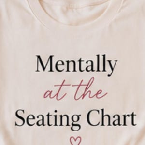 May include: Cream-colored t-shirt with the phrase "Mentally at the Seating Chart" in black and burgundy script, with a small heart symbol below. The shirt is folded, showcasing the design.
