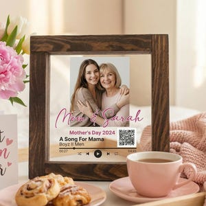 May include: A wooden framed photo with a picture of a mother and daughter, with the text "Mom & Sarah" and "Mother's Day 2024". A bouquet of pink peonies and white tulips, a card that says "Best Mom Ever", a cup of tea, and pastries are also in the image.