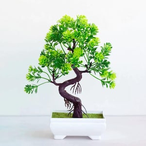 Pode incluir: Uma pequena árvore bonsai artificial em um vaso quadrado branco. A árvore tem um tronco e galhos marrom escuro com folhas verdes brilhantes. O vaso é preenchido com musgo verde.