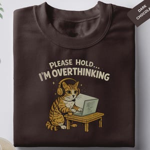 Puede incluir: Sudadera marrón chocolate oscuro con un gato de dibujos animados con auriculares y trabajando en una computadora portátil. El texto "PLEASE HOLD... I'M OVERTHINKING" está impreso encima del gato. Una etiqueta "Dark Chocolate" está adjunta.