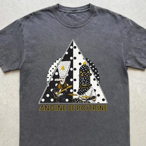 Puede incluir: Camiseta gris oscuro con un gráfico de dos figuras tocando instrumentos dentro de un triángulo con lunares. El texto "ANGINE DE POITRINE" está impreso en amarillo debajo. La camiseta tiene cuello redondo y mangas cortas.