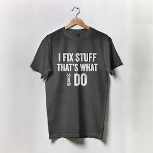 Może przedstawiać: Ciemnoszara koszulka z białym napisem "I FIX STUFF THAT'S WHAT I DO". Poniżej tekstu znajduje się grafika klucza. Koszulka wisi na drewnianym wieszaku na białym tle.