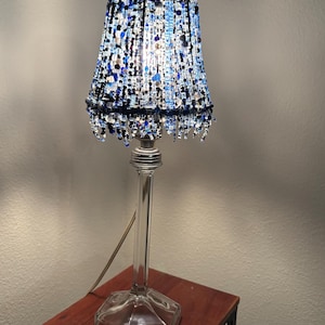 May include: A decorative table lamp with a clear glass base and a beaded lampshade. The lampshade is adorned with blue and clear beads, creating a textured and shimmering effect. The lamp is placed on a small, dark wooden table.