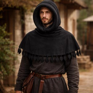 May include: A black hooded cloak with a decorative leaf-shaped trim. The cloak is worn over a dark gray tunic and a brown leather belt with a pouch. The person is standing outdoors.