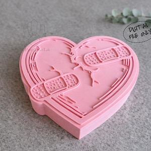 Puede incluir: Un cortador de galletas rosa en forma de corazón con un diseño de corazón roto, con dos curitas. El texto "DIGITAL FILE ONLY" está en un círculo punteado. El cortador está sobre una superficie gris.