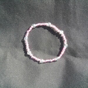 May include: A delicate bracelet featuring a pink cord strung with small, clear beads. Several larger, translucent beads are interspersed throughout the design. The bracelet is circular and rests on a dark, textured surface.