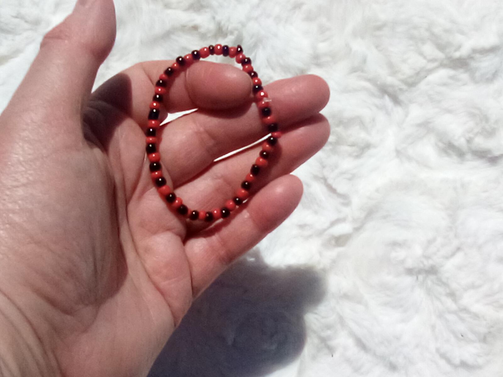 May include: A beaded bracelet featuring alternating red and black beads. The bracelet is made of small, round beads strung together to form a flexible band. The bracelet is shown against a white, fluffy background.