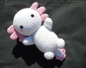Könnte beinhalten: Ein weißes, gehäkeltes Axolotl-Plüschtier mit rosa Kiemen und schwarzen Augen. Das Spielzeug liegt auf einer dunkelgrünen Oberfläche.