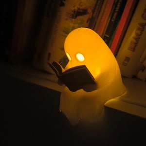 Peut inclure: Une veilleuse en forme de fantôme jaune lisant un livre. La lumière a deux découpes en forme d'yeux et un petit livre dans ses mains. La lumière est posée sur une étagère devant un fond flou de livres.