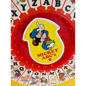 Pode incluir: Um brinquedo educativo circular com um painel central amarelo ilustrando o Mickey Mouse usando um capelo e segurando um livro. O texto diz MICKEY ABC'S. A borda vermelha ao redor exibe as letras do alfabeto.