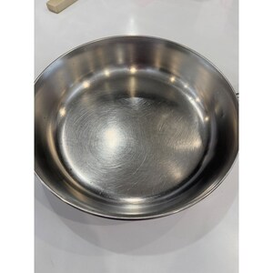 May include: A round, silver-coloured stainless steel frying pan with a smooth, reflective interior surface and a slightly raised rim. The metal shows subtle circular polishing marks across the base, indicating a durable, polished finish for cooking.