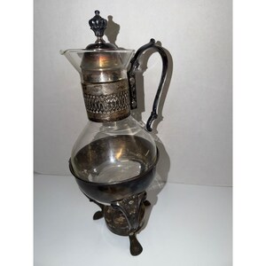 May include: A vintage silver and glass coffee maker. The glass carafe is held in place by a decorative silver metal frame. The coffee maker has a handle and a decorative lid. The base has three ornate feet.