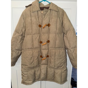 May include: A tan quilted puffer coat featuring a classic collar, long sleeves, and two large front patch pockets. The coat closes with three wooden toggle fasteners attached to brown leather-like tabs, creating a distinct front closure.