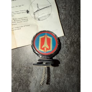 Puede incluir: Un adorno de capó circular cromado con un emblema central dorado sobre fondo azul, rodeado por un anillo rojo. El adorno tiene una base con resorte y se acompaña de una hoja de instrucciones con un diagrama que muestra la instalación.