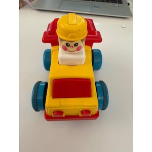 May include: A colorful toy dump truck with a yellow cab, red bed, and blue wheels. A yellow-helmeted figure sits in the cab. The toy is designed for young children and features a friendly face.