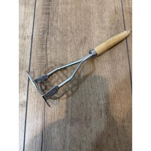 May include: A stainless steel potato masher with a wooden handle. The masher has a rectangular head with a grid pattern and a long handle for easy use. The handle is made of light-colored wood.