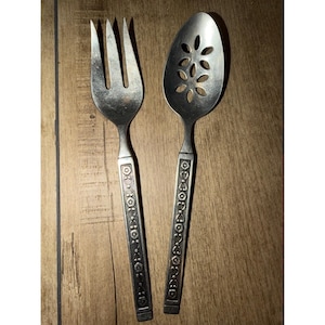 May include: A vintage silver-coloured serving fork and spoon set. The fork has three prongs, and the spoon features a slotted design with floral cutouts. Both utensils have decorative handles with an embossed floral pattern.